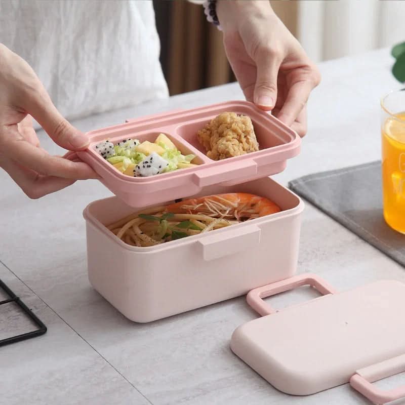 Pink bento box with compartments containing food, held by a person over a table with a glass of orange juice.