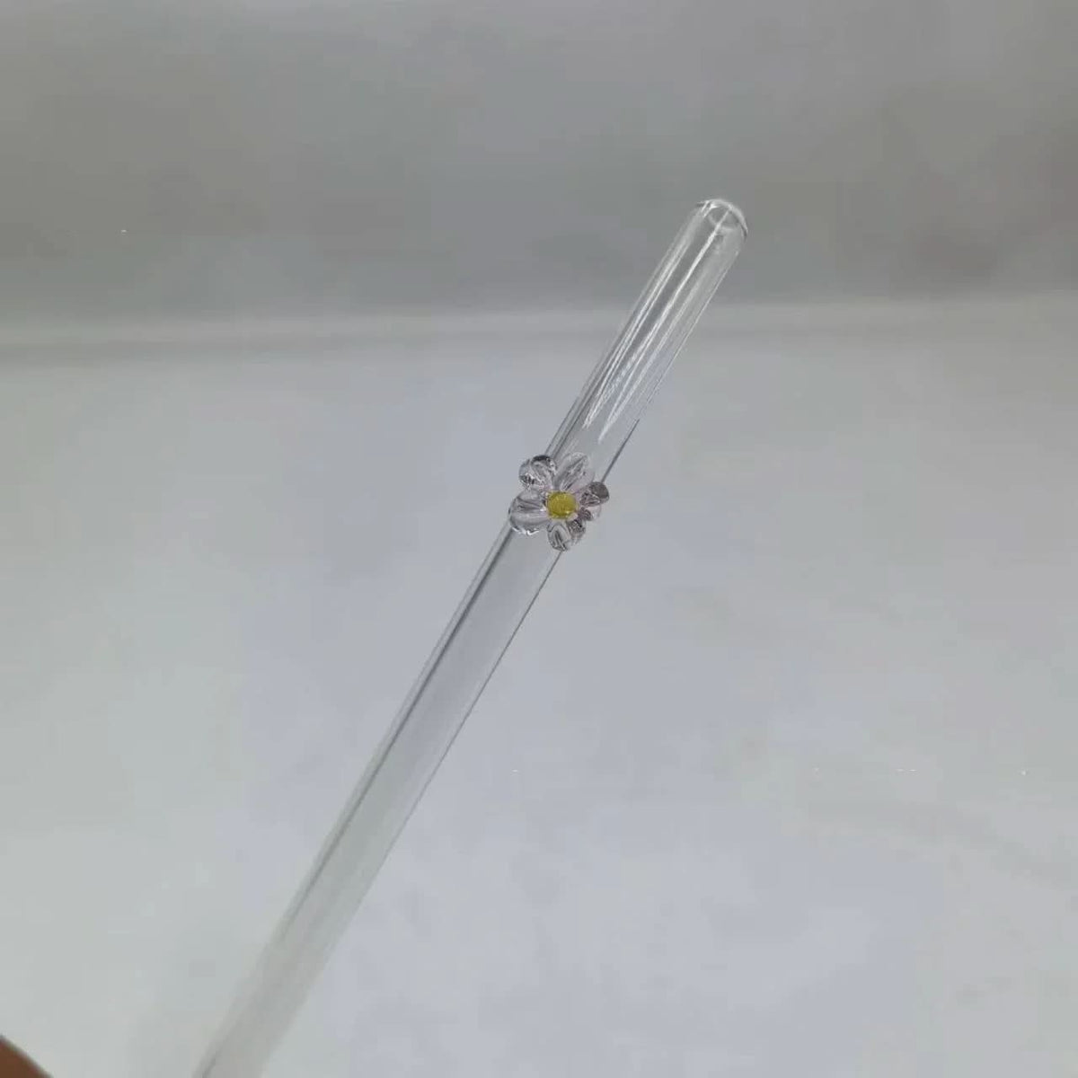 A clear glass straw with a small white and yellow daisy flower decoration near the top, shown against a plain light gray background.