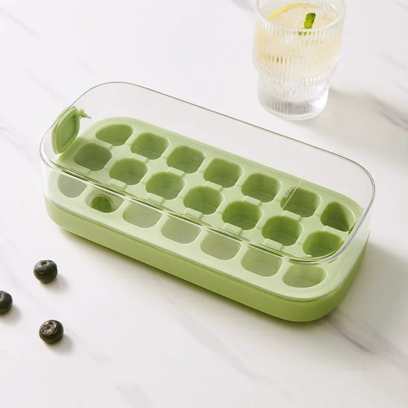 Green ice cube tray on a white surface with a glass of lemon water in the background.