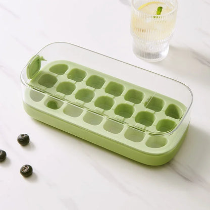 Green ice cube tray on a white surface with a glass of lemon water in the background.