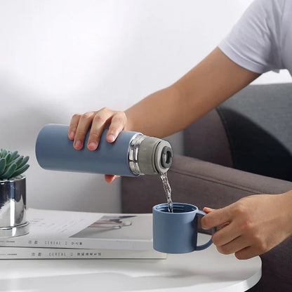 Person pouring liquid from a blue thermos into a matching mug on a table.