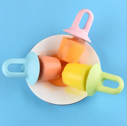 Three colorful silicone popsicle molds in light blue, light green, and light pink, shown with orange frozen popsicles on a white plate against a bright blue background.