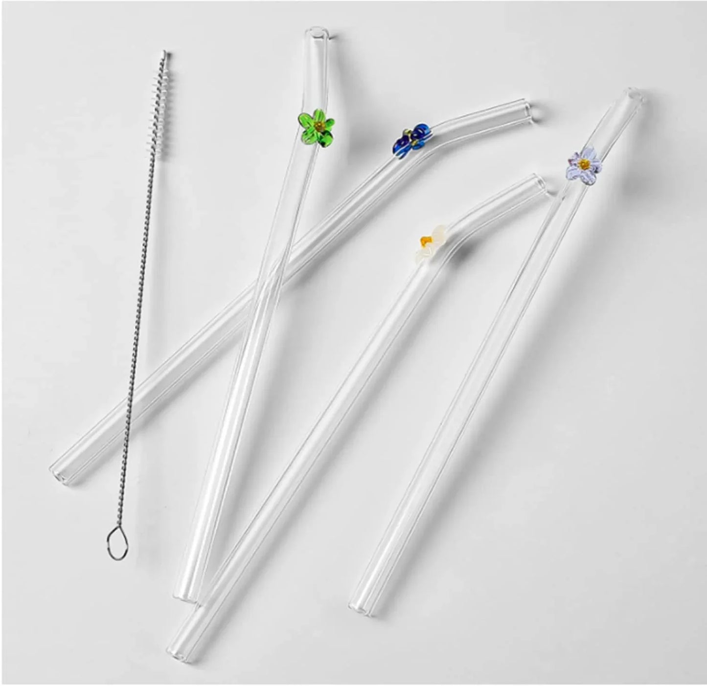 Four clear glass straws with small floral and leaf accents in green, blue, yellow, and white, plus a metal cleaning brush, arranged on a light gray background.
