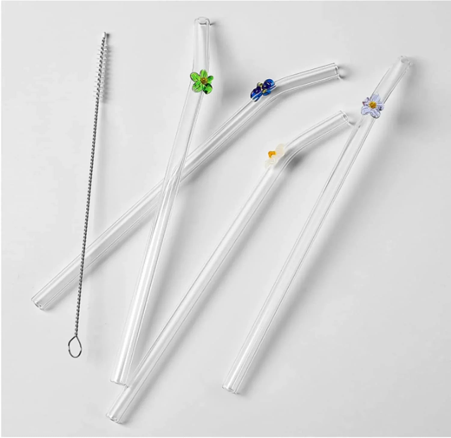 Four clear glass straws with small floral and leaf accents in green, blue, yellow, and white, plus a metal cleaning brush, arranged on a light gray background.