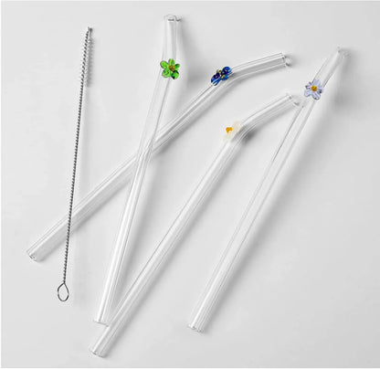 Four clear glass straws with small floral and leaf accents in green, blue, yellow, and white, plus a metal cleaning brush, arranged on a light gray background.