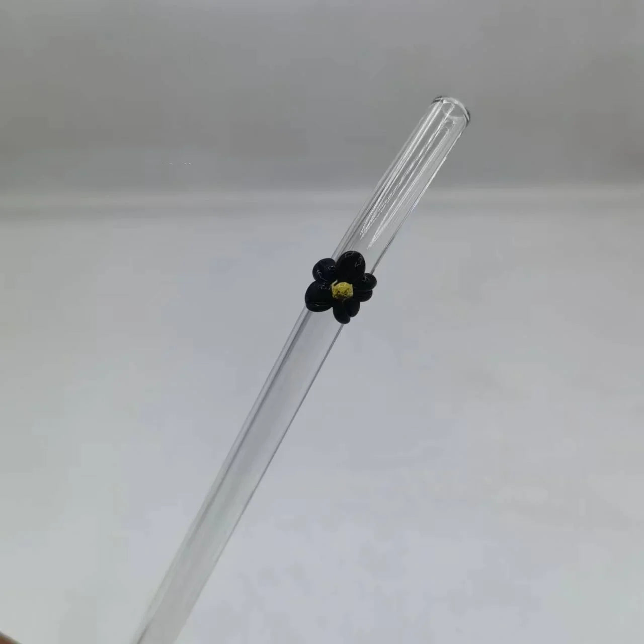 A clear glass drinking straw with a small black flower accent and a yellow center, shown against a plain light gray background.