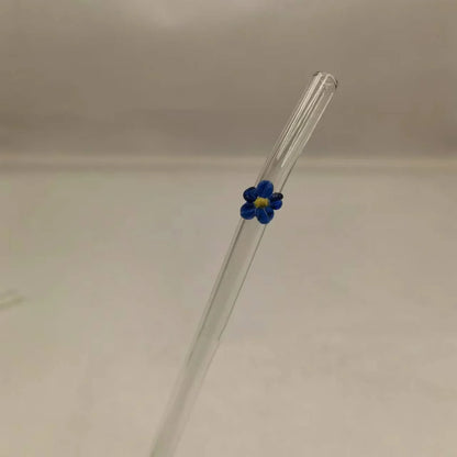 A clear glass straw with a small blue flower accent featuring a yellow center, set against a plain light gray background.
