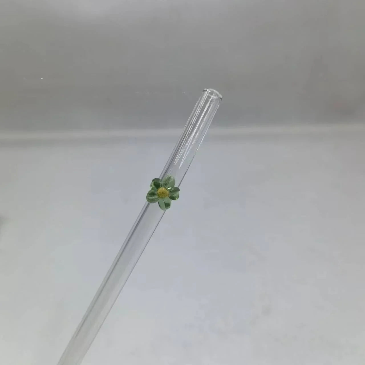 A clear glass drinking straw with a small green and yellow flower decoration near the top, shown against a plain light gray background.