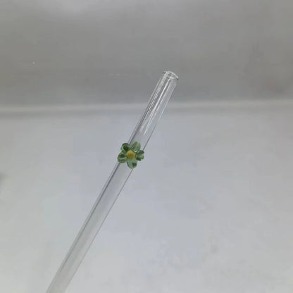 A clear glass drinking straw with a small green and yellow flower decoration near the top, shown against a plain light gray background.