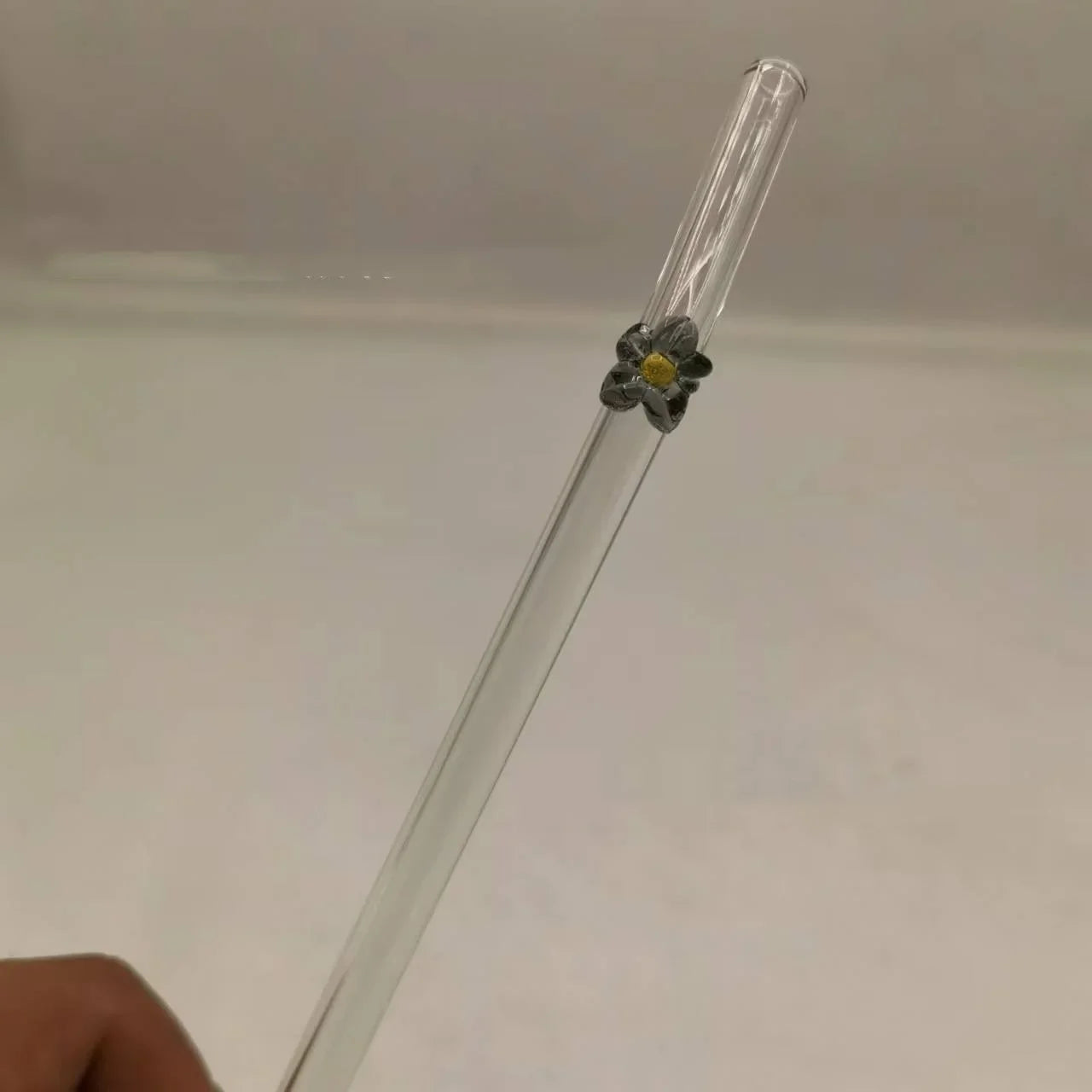 A clear glass drinking straw with a small gray flower accent and yellow center, held by a hand against a plain light background.
