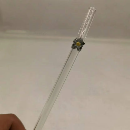A clear glass drinking straw with a small gray flower accent and yellow center, held by a hand against a plain light background.
