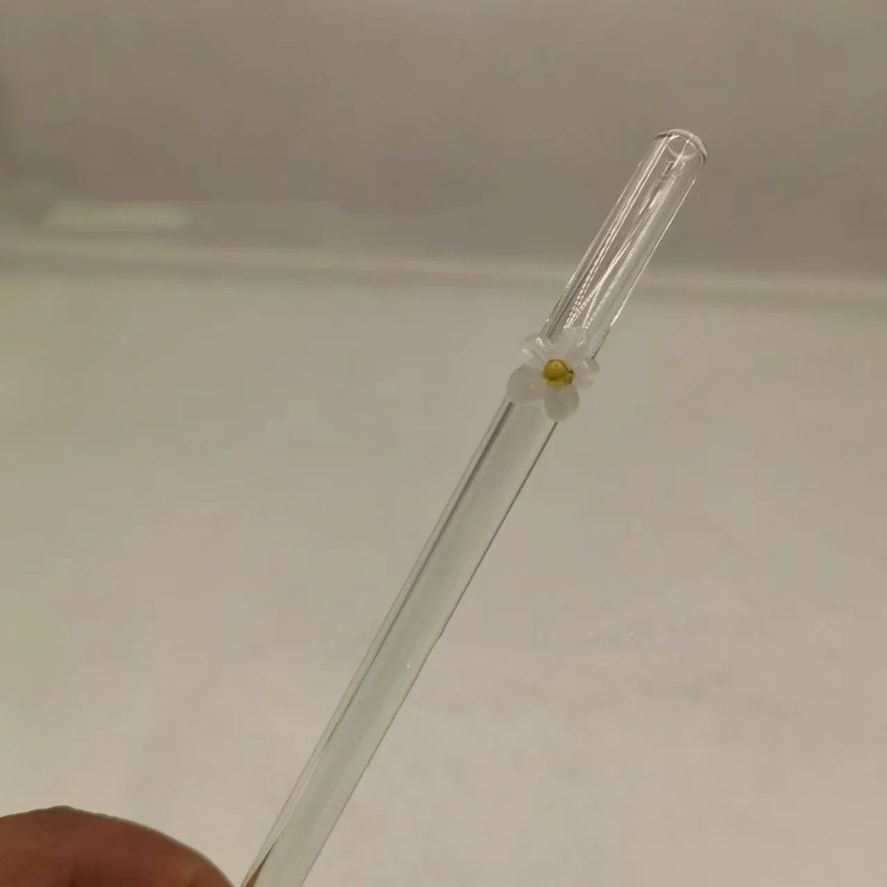 A clear glass drinking straw with a small white flower accent and yellow center, held by a hand against a plain light background.