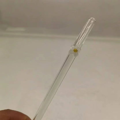 A clear glass drinking straw with a small white flower accent and yellow center, held by a hand against a plain light background.