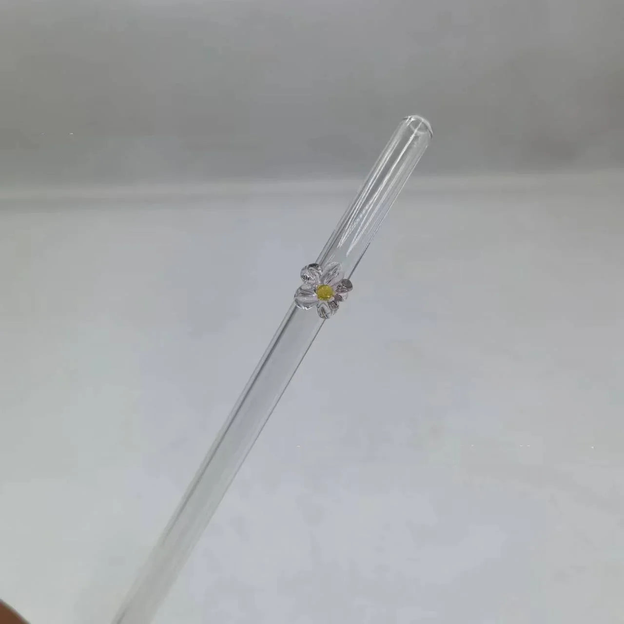 A clear glass straw with a small white and yellow daisy flower decoration near the top, shown against a plain light gray background.