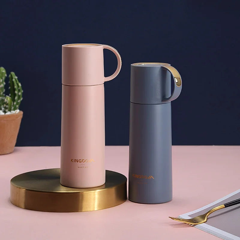 Two insulated travel mugs, one pink and one gray, on a pink surface with a dark blue background.