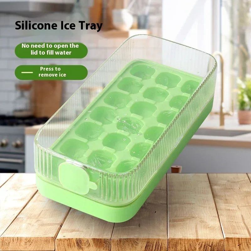A rectangular silicone ice tray in light green sits on a wooden countertop in a kitchen setting. The clear lid is slightly open, and the tray has multiple rounded ice cube compartments.